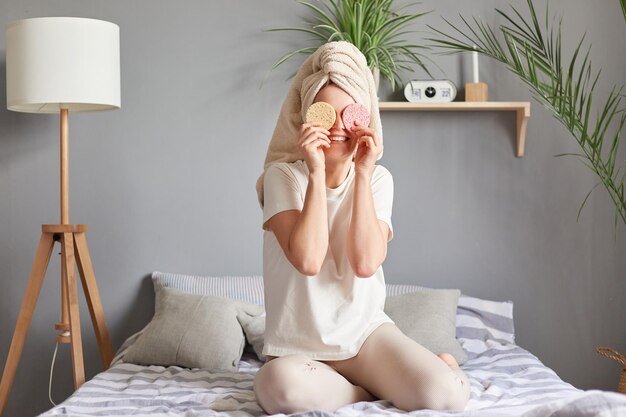 Fröhliche junge kaukasische Frau mit Handtuch auf dem Kopf, die zu Hause im Bett eine Hautpflegebehandlung durchführt und neue Schwämme hält, die ihre Augen bedecken, glücklich lächelt