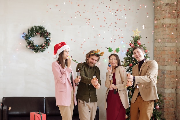 Fröhliche junge Kollegen in Weihnachtskopfbedeckungen schießen Cracker und amüsieren sich auf der Firmenfeier im Büro gegen Kranz an Wand und Couch