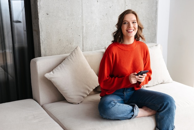 Fröhliche junge lässig gekleidete Frau, die auf einer Couch zu Hause sitzt und Handy hält