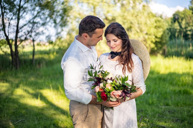 Fröhliche junge Leute in Hochzeitskleidern stehen auf der Wiese und schauen sich gegenseitig an Porträt einer Braut, die einen schönen Hochzeitsstrauß ansieht und ihr Mann seine Frau ansieht