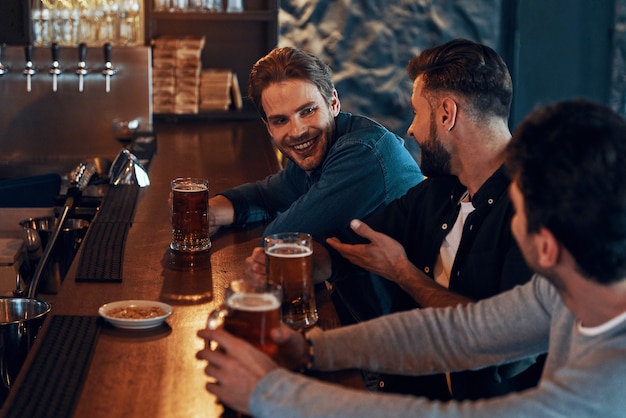 Fröhliche junge Männer in Freizeitkleidung trinken Bier und verbinden sich beim Sitzen in der Kneipe