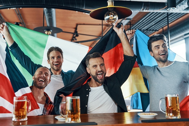 Fröhliche junge Männer mit internationalen Flaggen, die Bier trinken und Sportspiele sehen, während sie in der Kneipe sitzen?