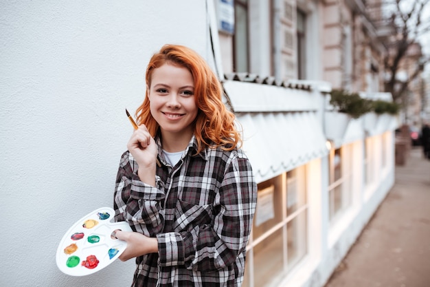 Fröhliche junge Malerin mit roten Haaren, die auf die Straße geht, während sie Palette und Pinsel im Freien hält.