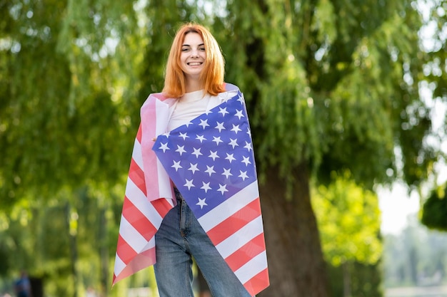 Fröhliche junge rothaarige Frau, die mit der US-Nationalflagge auf ihren Schultern posiert und draußen im Sommerpark steht Hübsches Mädchen, das den Unabhängigkeitstag der Vereinigten Staaten feiert