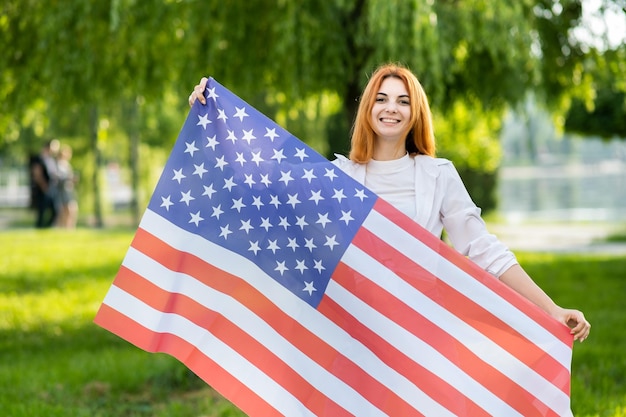 Fröhliche junge rothaarige Frau, die mit der US-Nationalflagge posiert, die draußen im Sommerpark steht Positives Mädchen mit Banner der Vereinigten Staaten im Freien