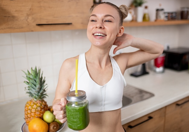 Fröhliche junge schlanke Frau in der weißen Spitze, die Glastopf mit frisch gemachtem gesundem Detox-grünem Smoothie hält, während in der Küche steht