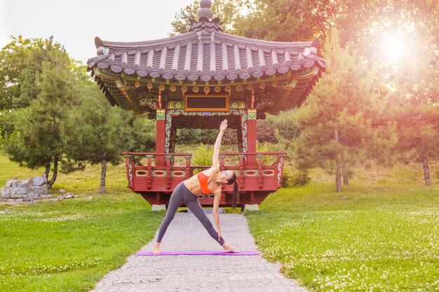 Fröhliche junge schöne Frau, die Yoga im ostasiatischen Park praktiziert und aufwirft. Dreieckshaltung.