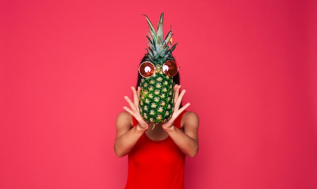 Fröhliche junge schöne Frau in roter Badeanzug-Sonnenbrille und großem Sommerhut mit Ananas in Sonnenbrille in der Hand posiert auf rosa Hintergrund