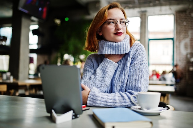 Fröhliche junge schöne rothaarige Frau mit Brille, die ihr Telefon-Touchpad und Notizbuch benutzt, während sie an ihrem Arbeitsplatz im Café mit einer Tasse Kaffee sitzt