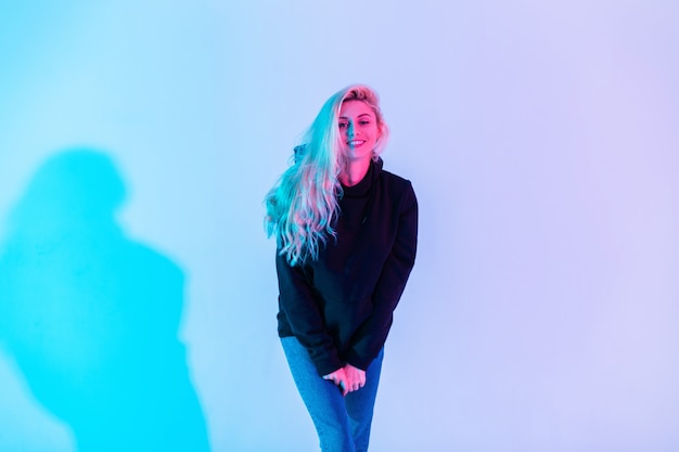 Fröhliche junge Schönheit süße Frau mit blonden Haaren und weißem Lächeln in modischem schwarzen Hoodie und blauen Jeans auf einem farbigen neonrosa Licht im Studio