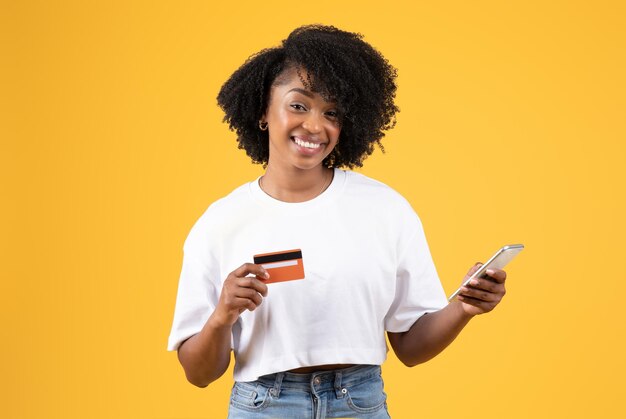 Fröhliche junge schwarze, lockige Dame im weißen T-Shirt zeigt Kreditkarte und Smartphone isoliert auf Orange