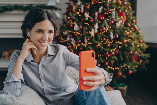 Fröhliche junge Spanierin genießt die Weihnachtszeit zu Hause und macht Selfie auf dem Smartphone