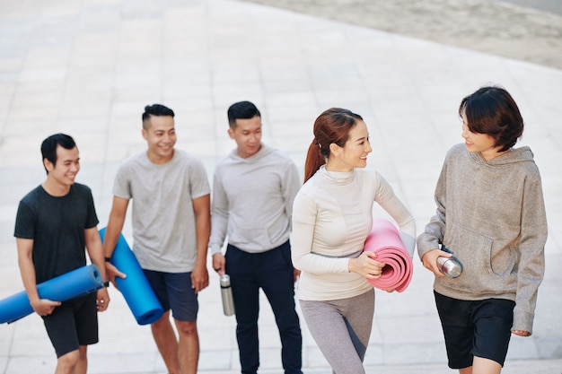 Fröhliche junge, sportliche Vietnamesen mit Yogamatten, die nach dem Training im Freien nach Hause gehen