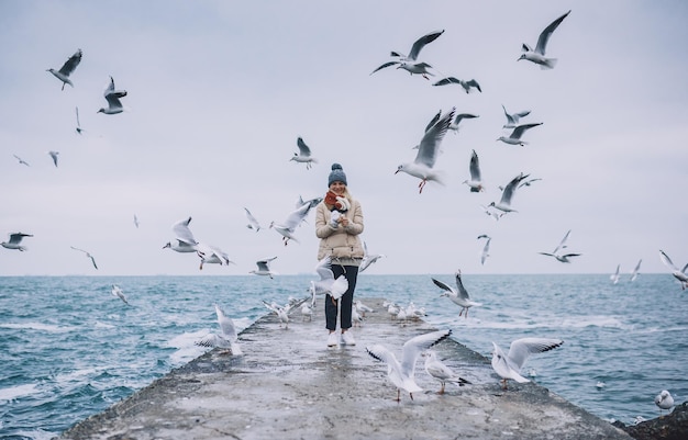 Fröhliche junge Touristenfrau füttert Möwen auf dem Meer Hübsche Frau, die Mantelschal trägt und fliegende Möwen am Meer am Himmel beobachtet Menschen reisen Umwelt Naturkonzept