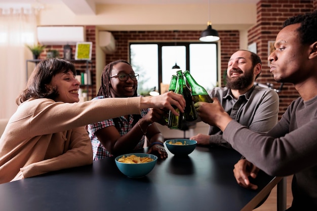 Fröhliche junge, vielfältige Menschen sitzen am Tisch und entspannen sich bei Bier und Snacks. Positive multiethnische Gruppe von Freunden, die zu Hause im Wohnzimmer gemeinsam trinken und Spaß haben.