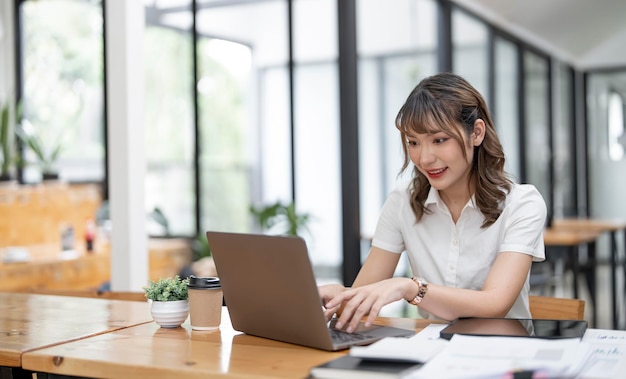 Fröhliche junge weibliche Geschäftsfrau oder Studentin, die im Büro am Computer-Laptop lächelt und arbeitet