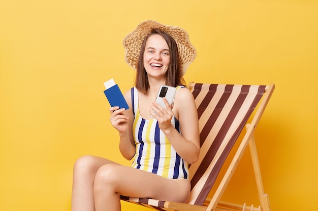 Fröhliche kaukasische erwachsene Frau in gestreiftem Badeanzug sitzt auf Liegestuhl isoliert über gelbem Hintergrund mit Handy, das Passbuchungstickets im Ausland hält