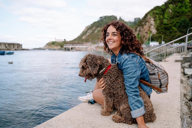 Fröhliche kaukasische Frau, die ihren Wasserhund über den Hafenweg umarmt. Horizontale Ansicht der Frau, die mit Tier reist. Tiere und Reisekonzept.