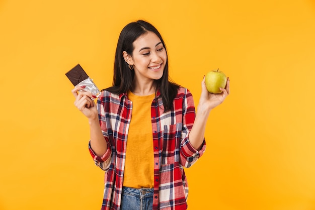 fröhliche kaukasische Frau, die lächelt, während sie Apfel und Schokolade isoliert über gelber Wand hält?
