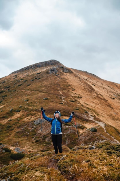 Fröhliche kaukasische Frau in blauer Jacke mit modernen Trekkingstäbchen in den Händen steht in den hohen Bergen auf dem Gipfel von Hiking Freizeitaktivität und Menschenkonzept