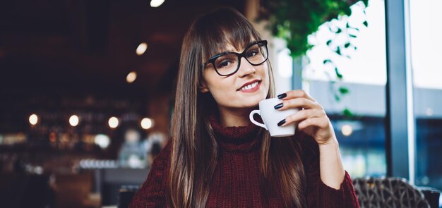 fröhliche kaukasische frau in trendigen brillen zur sehkorrektur, die eine tasse mit heißem tee hält