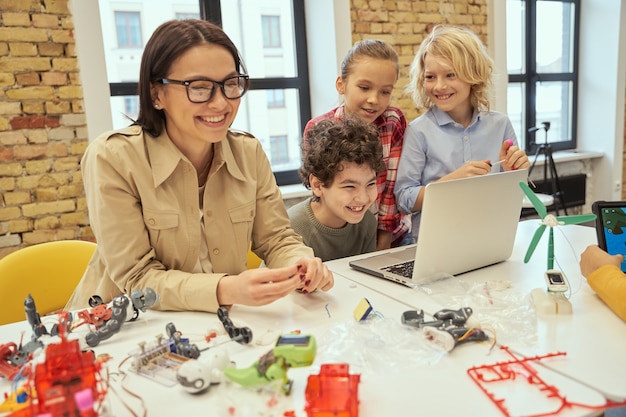 Fröhliche Kinder lachen zusammen mit ihrer jungen Lehrerin, während sie lernen, wie man Roboter zusammenbaut