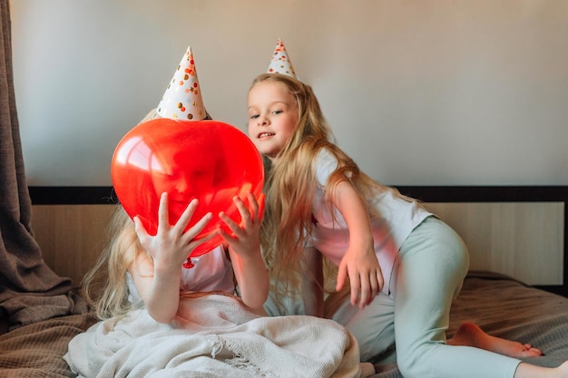 Fröhliche Kinder Mädchen Schwestern Zwillinge zu Hause auf dem Bett morgens an ihrem Geburtstag in festlichen Mützen mit einem roten Ballon in Form eines Herzens