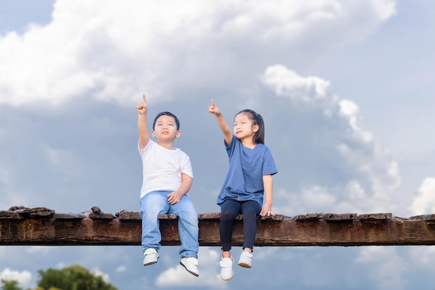 Fröhliche Kinder mit Beschneidungspfad sitzen auf einer Holzbrücke Fröhliche Kinder, Junge und Mädchen, die Spaß im Freien haben, Bruder und Schwester, die im Garten mit verschwommenem Himmelshintergrund spielen