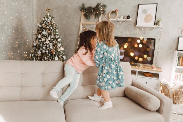 Fröhliche kleine Kinder, kleine Mädchen springen lachend auf die Couch und haben Spaß beim gemeinsamen Spielen zu Hause, Kinder genießen unbeschwerte Freizeit zu Hause