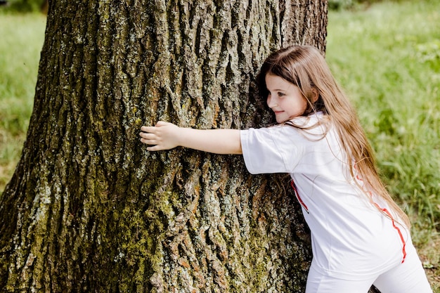 Fröhliche kleine Mädchen umarmen einen Baumstamm im Park im Freien