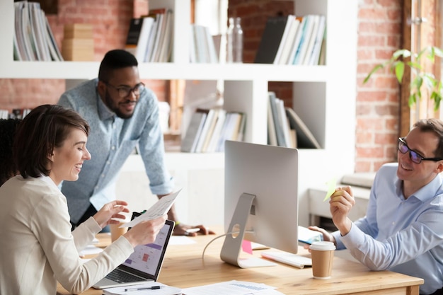 Fröhliche Kollegen arbeiten an Computern in Coworking-Büroräumen und diskutieren lachend über Papierkram-Projekte
