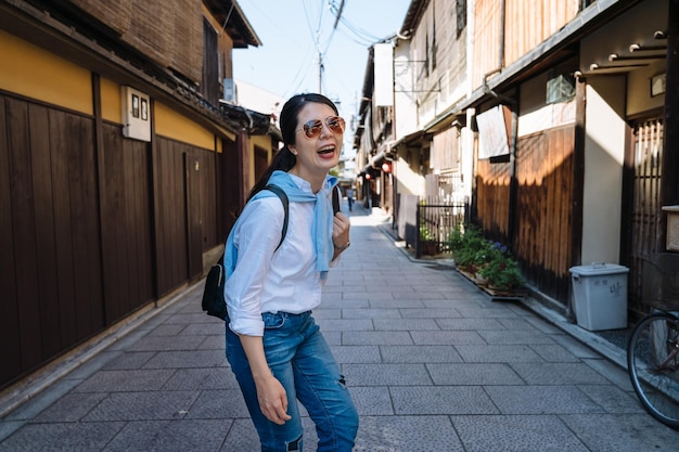 Fröhliche Koreanerin mit Sonnenbrille dreht sich um und blickt mit einem breiten Lächeln in die Kamera, während sie Spaß am Urlaub hat, indem sie die Hanamikoji-Straße in Gion, Japan, besucht.