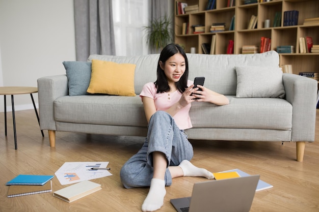 Fröhliche koreanische Dame entspannt sich zu Hause mit Laptop und Smartphone im Wohnzimmer und sendet Nachrichten auf dem Handy