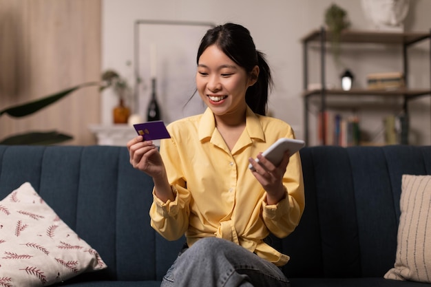 Fröhliche koreanische Dame mit Kreditkarte und Handy-Shopping drinnen