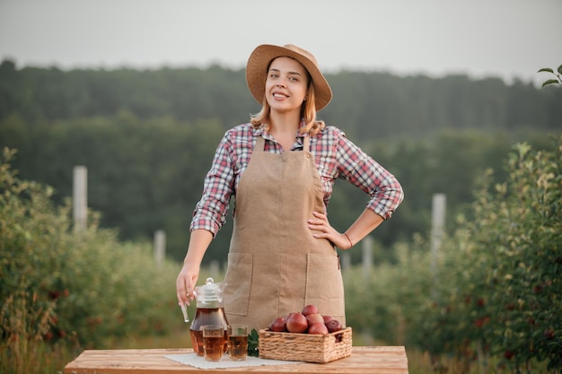 Fröhliche lächelnde Bäuerin mit leckerem Saft und frischen reifen Äpfeln im Obstgarten während der Herbsternte Erntezeit