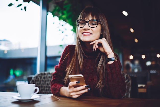 Fröhliche lächelnde Bloggerin, die sich in der Cafeteria ausruht und drinnen ein Smartphone-Gadget in der Hand hält