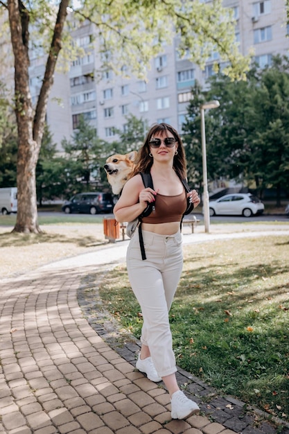 Fröhliche lächelnde Frau, die draußen in der Parklandschaft der Stadt mit dem Hund Welsh Corgi Pembroke in einem speziellen Rucksack spazieren geht