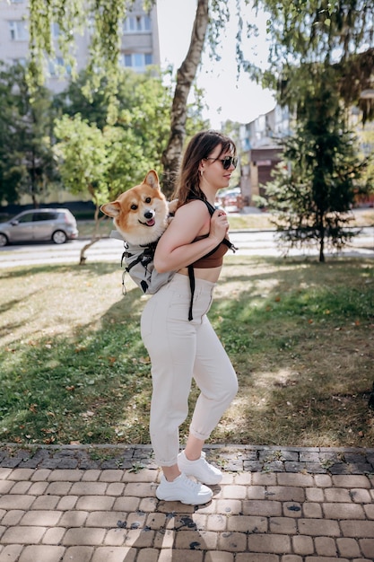 Fröhliche lächelnde Frau, die draußen in der Parklandschaft der Stadt mit dem Hund Welsh Corgi Pembroke in einem speziellen Rucksack spazieren geht
