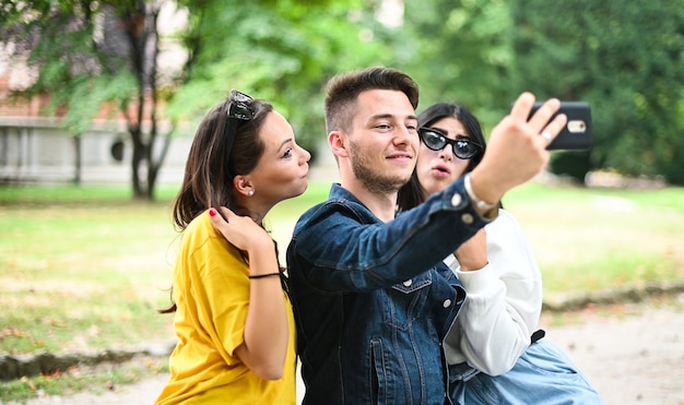 Fröhliche lächelnde Freunde im Park sitzen auf einer Bank und machen Selfies mit einem Smartphone