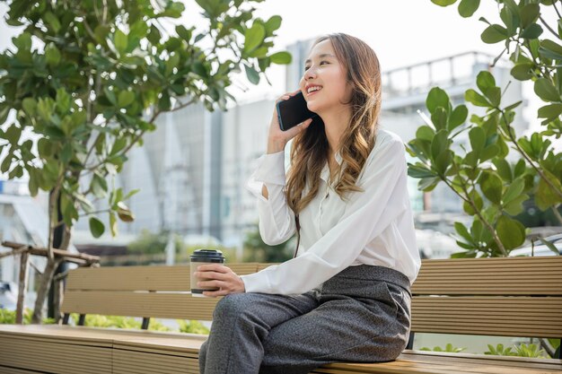 Fröhliche lächelnde Geschäftsfrau, die am Handy außerhalb der Straßenstadt in der Nähe des Büros sitzt, asiatische junge Geschäftsfrau, die ein mobiles Smartphone benutzt, das auf dem Außengebäude des Mobiltelefons im Freien spricht
