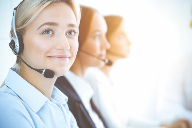 Fröhliche lächelnde Geschäftsfrau mit Kopfhörern, die Kunden beraten. Gruppe verschiedener Telefonisten bei der Arbeit im sonnigen Büro. Konzept für Call Center und Geschäftsleute.