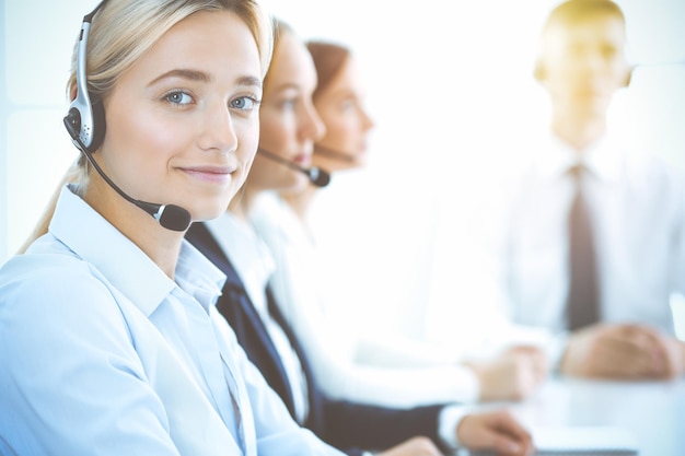 Fröhliche lächelnde Geschäftsfrau mit Kopfhörern, die Kunden beraten. Gruppe verschiedener Telefonisten bei der Arbeit im sonnigen Büro. Konzept für Call Center und Geschäftsleute.