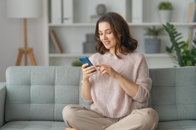 Fröhliche, lässige, schöne Frau benutzt ein Telefon, das zu Hause auf einem Sofa sitzt