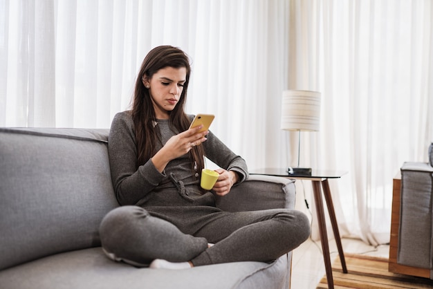 Fröhliche lateinamerikanische Dame entspannt sich beim Kaffeetrinken zu Hause allein sitzen auf der Zimmerpose und teilen gute Nachrichten