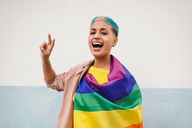 Fröhliche lesbische Frau mit LGBT-Regenbogenfahne, während sie vor der Kamera lächelt und Friedensgeste macht