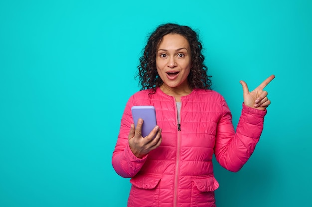 Fröhliche, lockige Brünette mit weit geöffneten Augen überrascht mit Blick auf die Kamera, die das Handy hält und mit dem Finger auf blauen Hintergrund mit Kopienraum zeigt