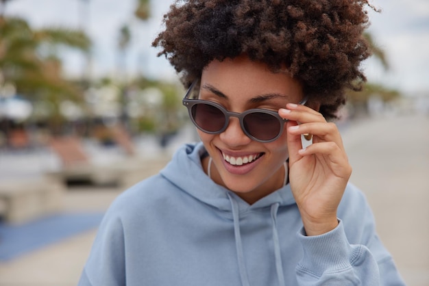 Fröhliche lockige Frau trägt stylische Sonnenbrillen und lässiges Hoodie-Lächeln verbringt glücklich Freizeit bei Straßenposen im Freien vor verschwommenem Hintergrund. Lebensstil der Menschen und Konzept der guten Laune