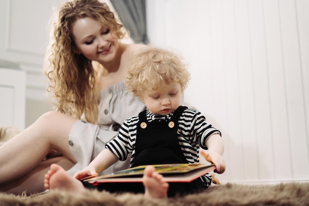 Fröhliche lockige junge Mutter, die mit ihrem kleinen Sohn auf dem Boden sitzt und ein Buch liest und Spaß hat