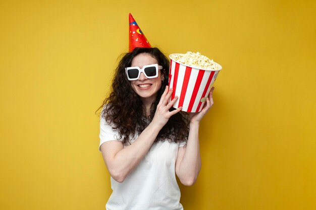 Fröhliche, lockige Zuschauerin mit 3D-Brille und festlichem Hut hält Popcorn in der Hand und feiert Geburtstag