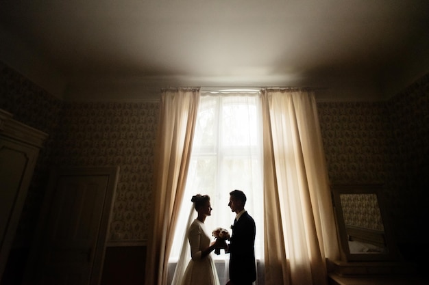 Fröhliche Luxusbraut und Bräutigam, die in einem zärtlichen Moment am Fensterlicht stehen